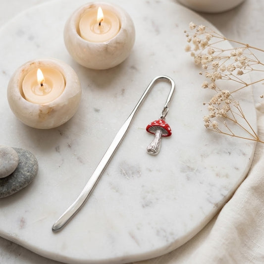 A silver metal bookmark shaped like a small red-capped mushroom with white spots, resting on a round white marble tray. The scene includes two lit candles, dried flowers, and smooth stones in a softly styled setting.