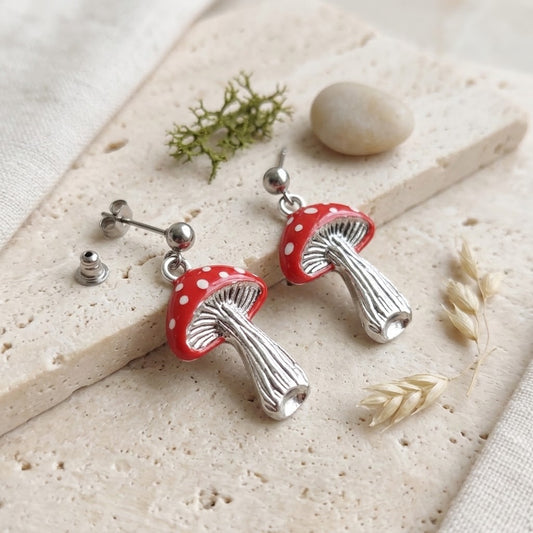 A pair of mushroom-shaped earrings with red caps dotted with white spots and textured silver stems, displayed on a beige stone slab with small dried botanicals.