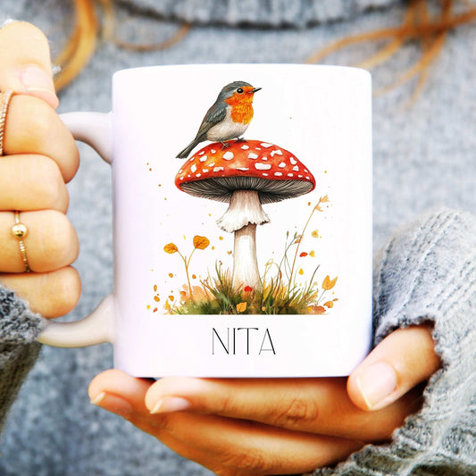Person holding a mug with a design of a robin on a mushroom, labelled 'NITA'.