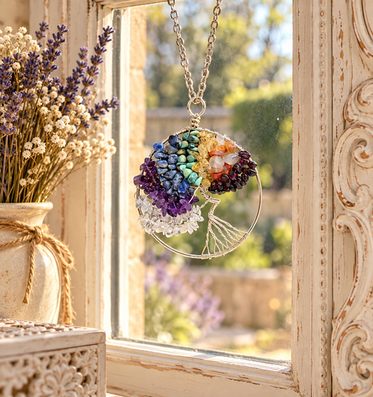 A circular chakra pendant necklace hanging in front of a sunlit window. The pendant features multiple coloured gemstone sections representing the chakras, with a silver-toned chain and circular frame.