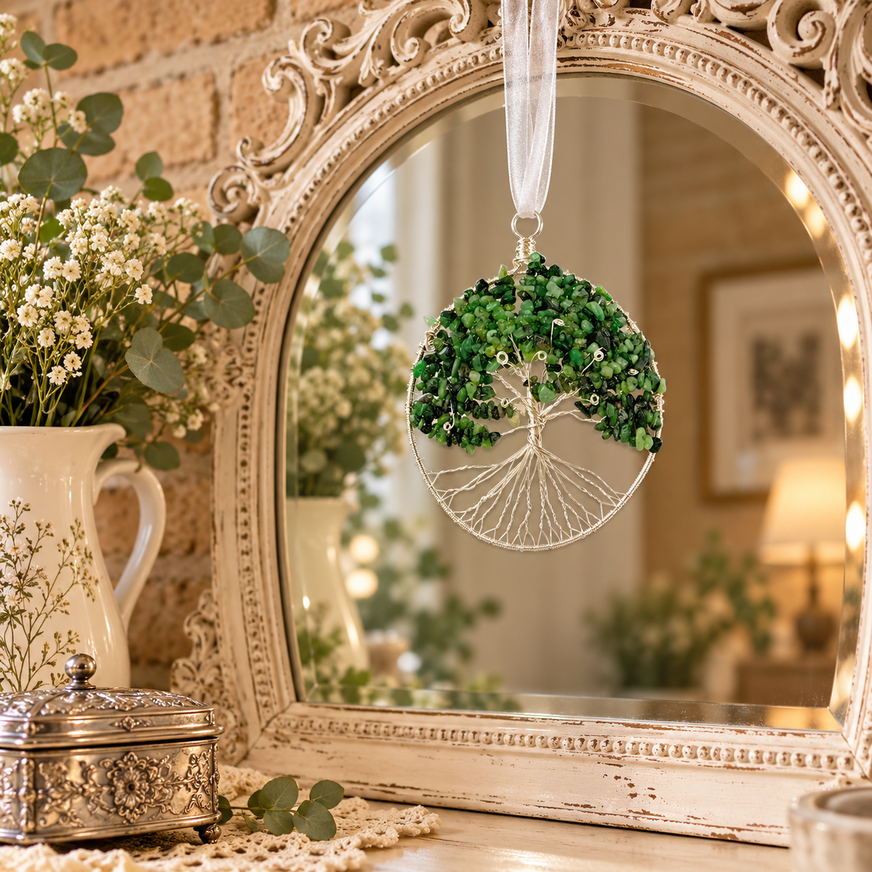 A decorative hanging ornament shaped like a tree with green leaves and a white wire trunk, suspended in front of an ornate cream mirror. The scene is styled with a white pitcher of white flowers, greenery, and warm indoor lighting in the background.