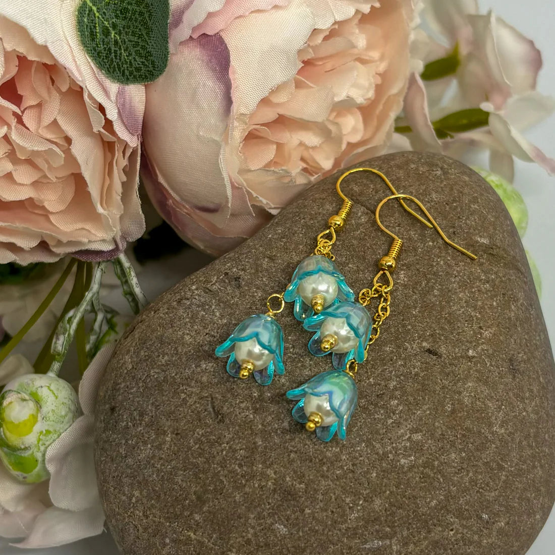 Gold and blue floral earrings on a stone with pink flowers in the background