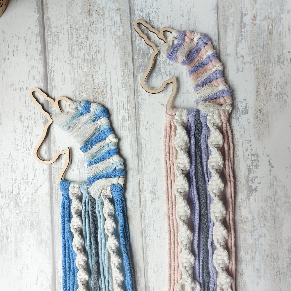 Two macrame unicorn wall hangings in blue and pink colours, made from braided cotton, displayed on a wooden background.