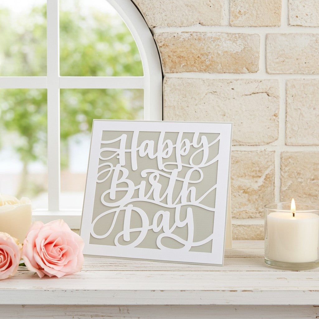 A square white unisex birthday card reading 'Happy Birthday' in elegant cursive, displayed on a white windowsill with pink roses, a lit candle in a glass holder, and a light stone brick wall background.