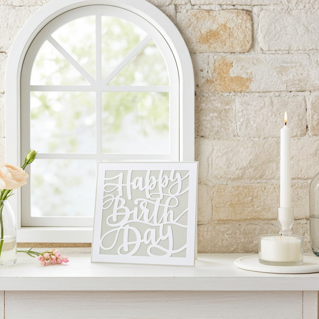 A white square greeting card with the words 'Happy Birthday' in a cursive cutout font, displayed on a white mantelpiece next to a lit white candle, a brick wall, and a window.