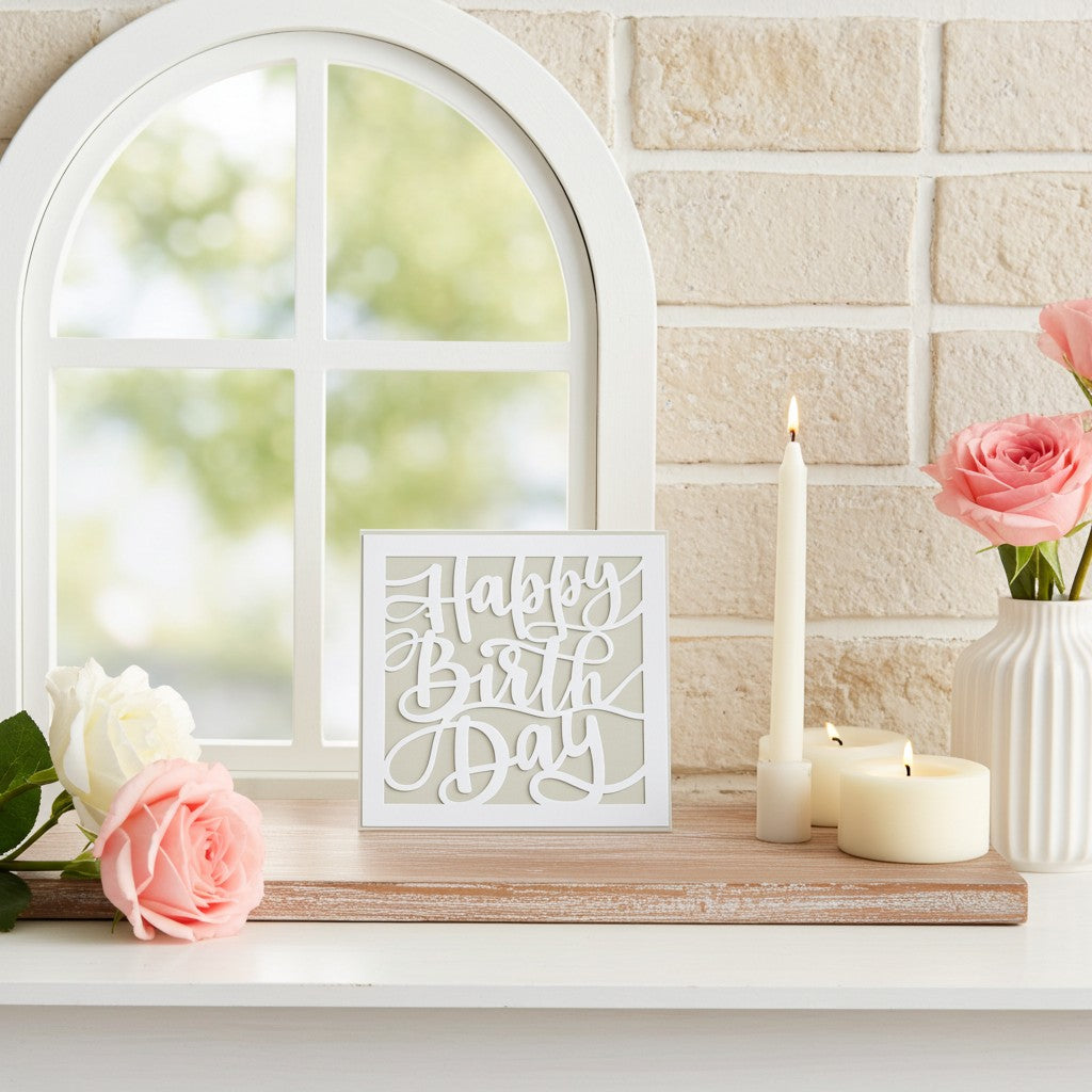 A white square unisex greeting card reading 'Happy Birthday' in cursive script, styled on a wooden tray on a white mantel next to pink and white roses, candles, and a white arched window frame against a beige brick wall.