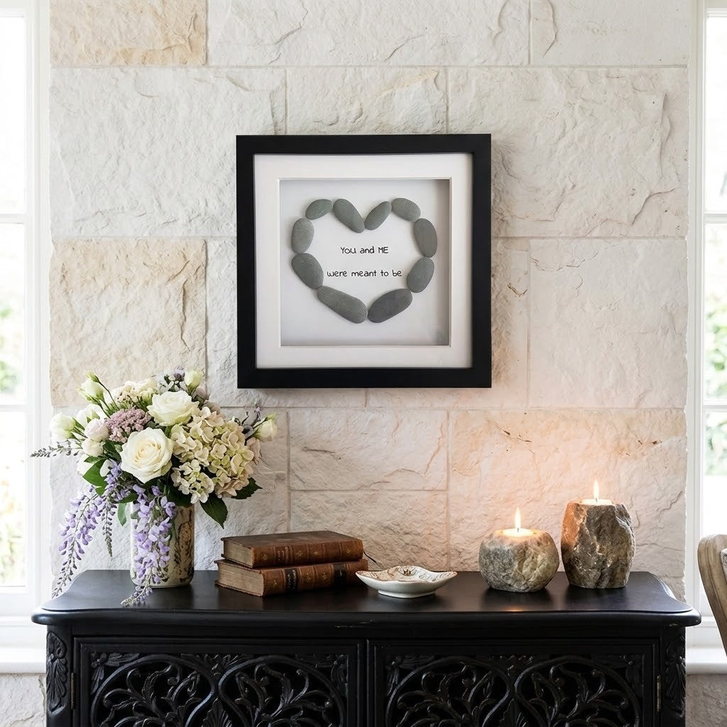 Framed shadow box wall art hanging on a light stone wall above a dark console table. The artwork shows grey stones arranged in a heart shape with the words 'you and I love meant to be' in the center.