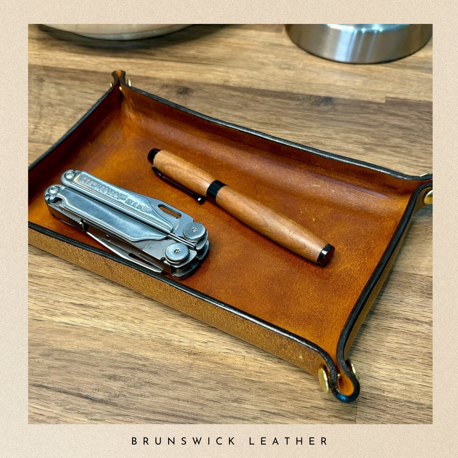 Brown leather tray with a multitool and pen on a wooden surface, branded 'Brunswick Leather'.