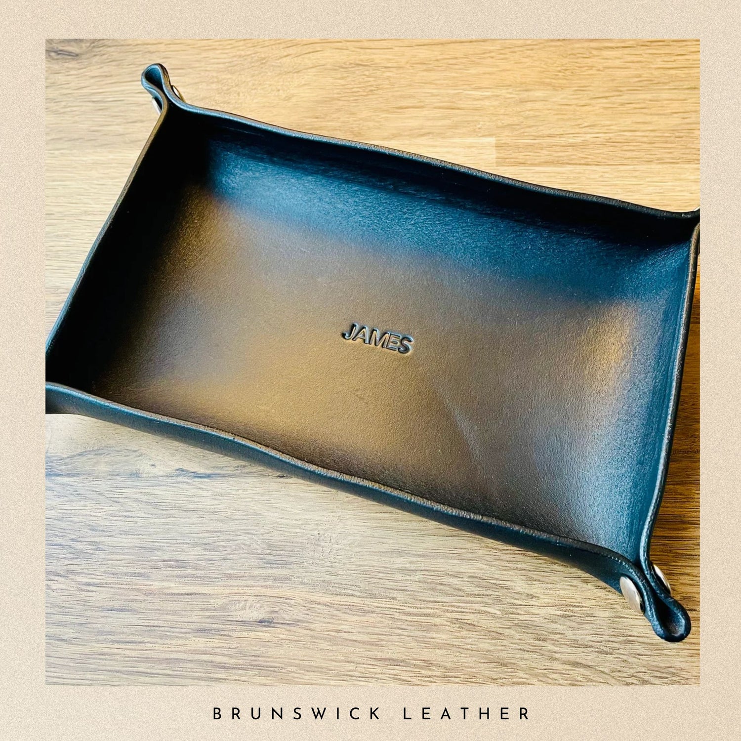Black leather tray with 'JAMES' branding on a wooden surface, featuring Brunswick Leather.