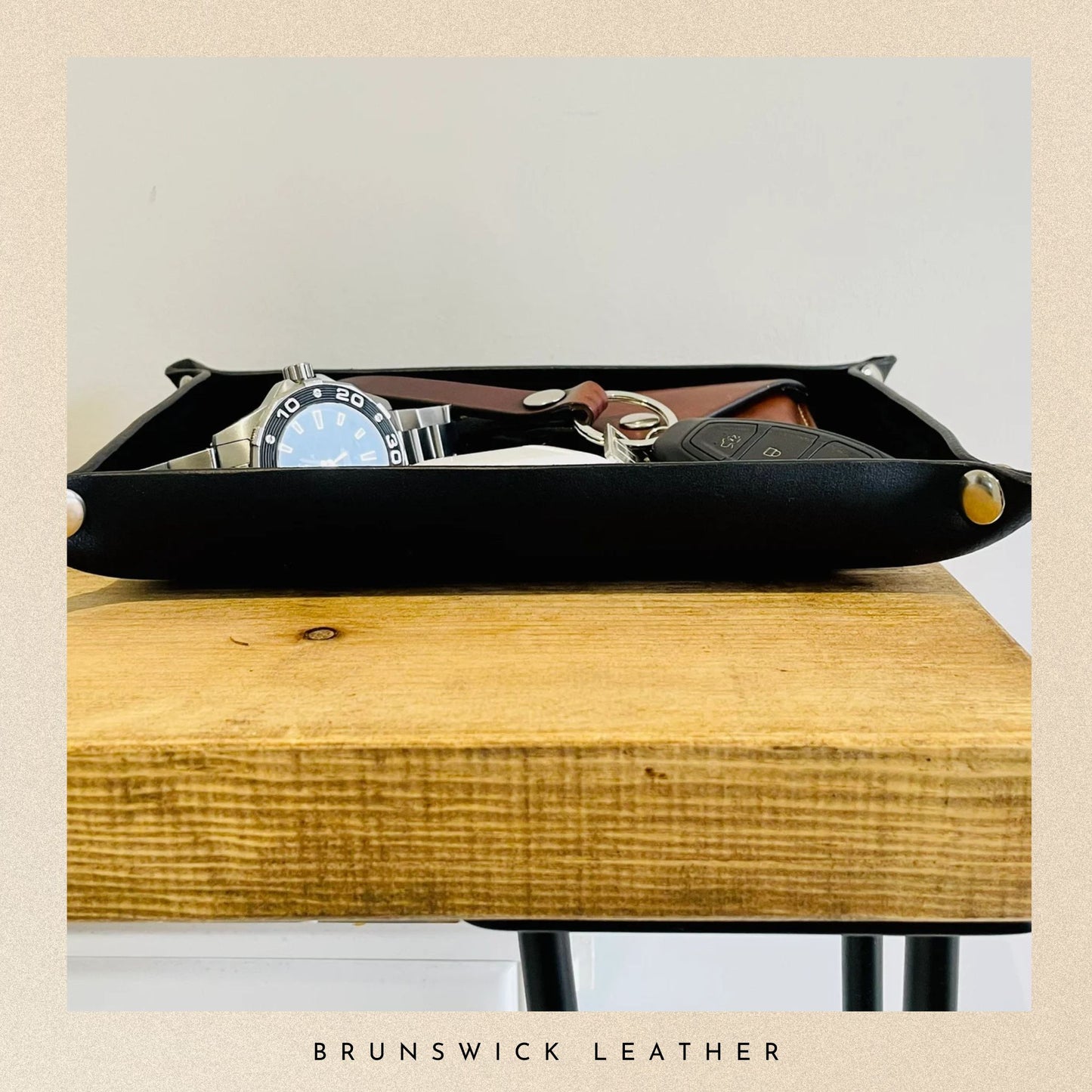 Black leather tray with watch, keys, and wallet on a wooden surface with 'Brunswick Leather' branding.