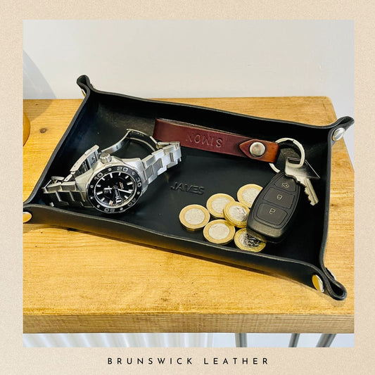 Black leather tray with watch, keys, and coins on a wooden surface, branded 'Brunswick Leather'.