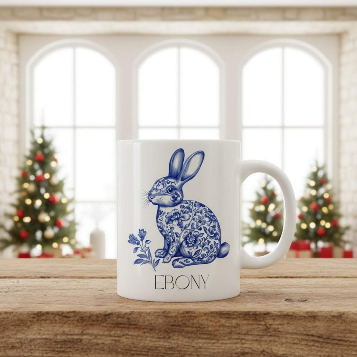 White mug with blue hare design and 'Ebony' text on a wooden surface with Christmas trees in the background.