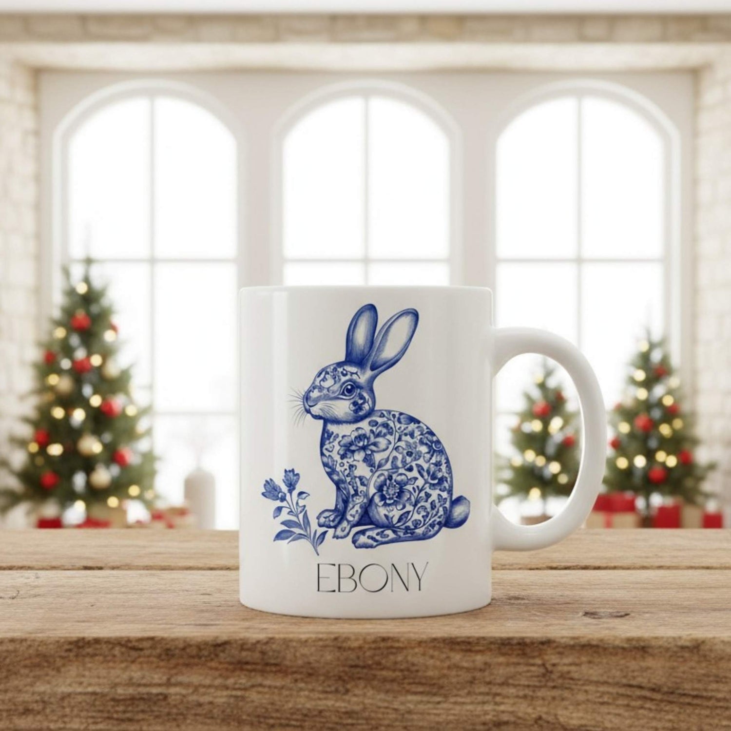 White mug with blue hare design and 'Ebony' text on a wooden surface with Christmas trees in the background.