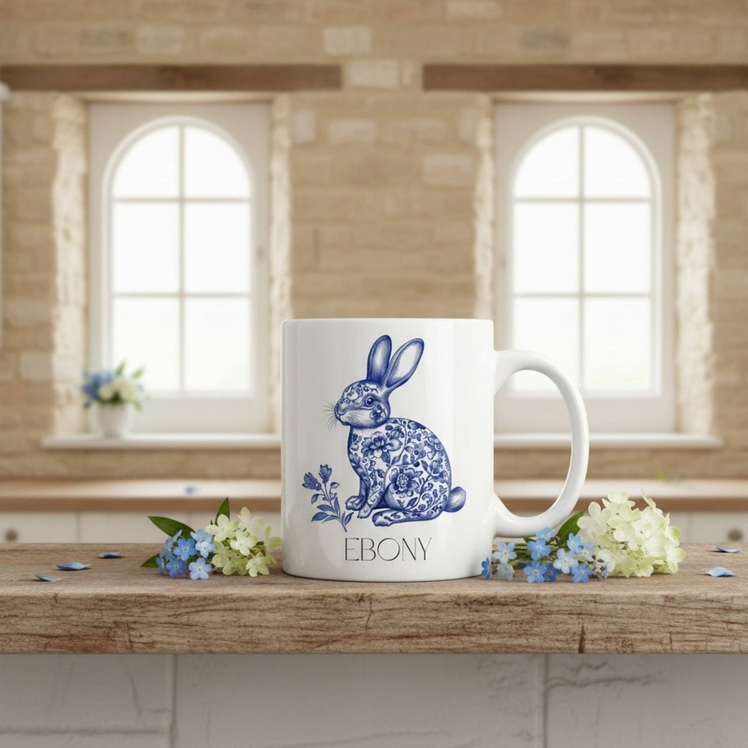 White mug with blue hare design and 'EBONY' text on a wooden surface with flowers, in a room with large windows.