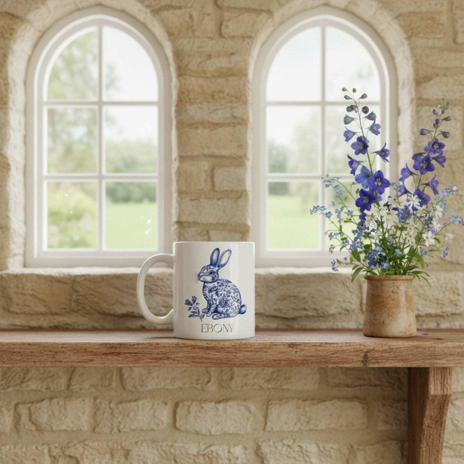 White mug with blue hare design on a wooden table with a stone wall and windows in the background