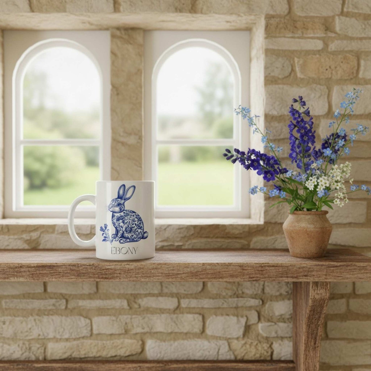 White mug with blue hare design on a wooden table in front of stone walls and windows.