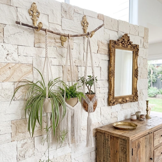 A boho-style macrame plant hanger with a woven basket pot holding a small green plant, hanging from a rustic wooden branch mounted on a light stone wall. The scene includes a gold mirror and a wooden cabinet, creating a warm, styled interior setting.