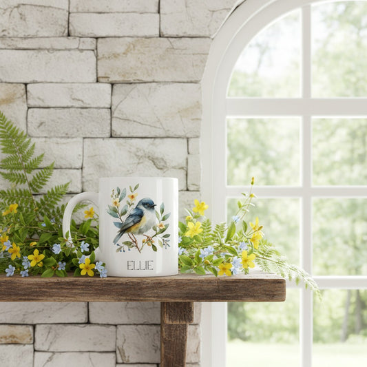 Mug with bird design and 'Ellie' text on a wooden surface with flowers and a stone wall background