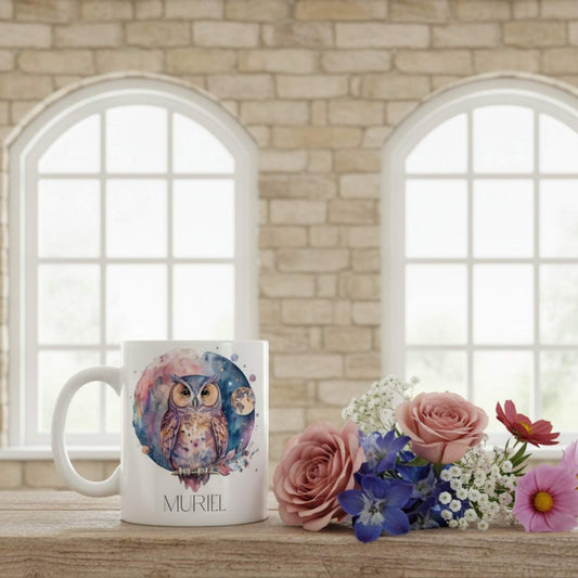Mug with owl design and name 'Muriel' on a wooden surface with flowers, against a stone wall and window background.