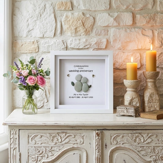 A framed wedding anniversary keepsake display with pebble arrangement, styled on a white cabinet against a stone wall with flowers and lit candles.