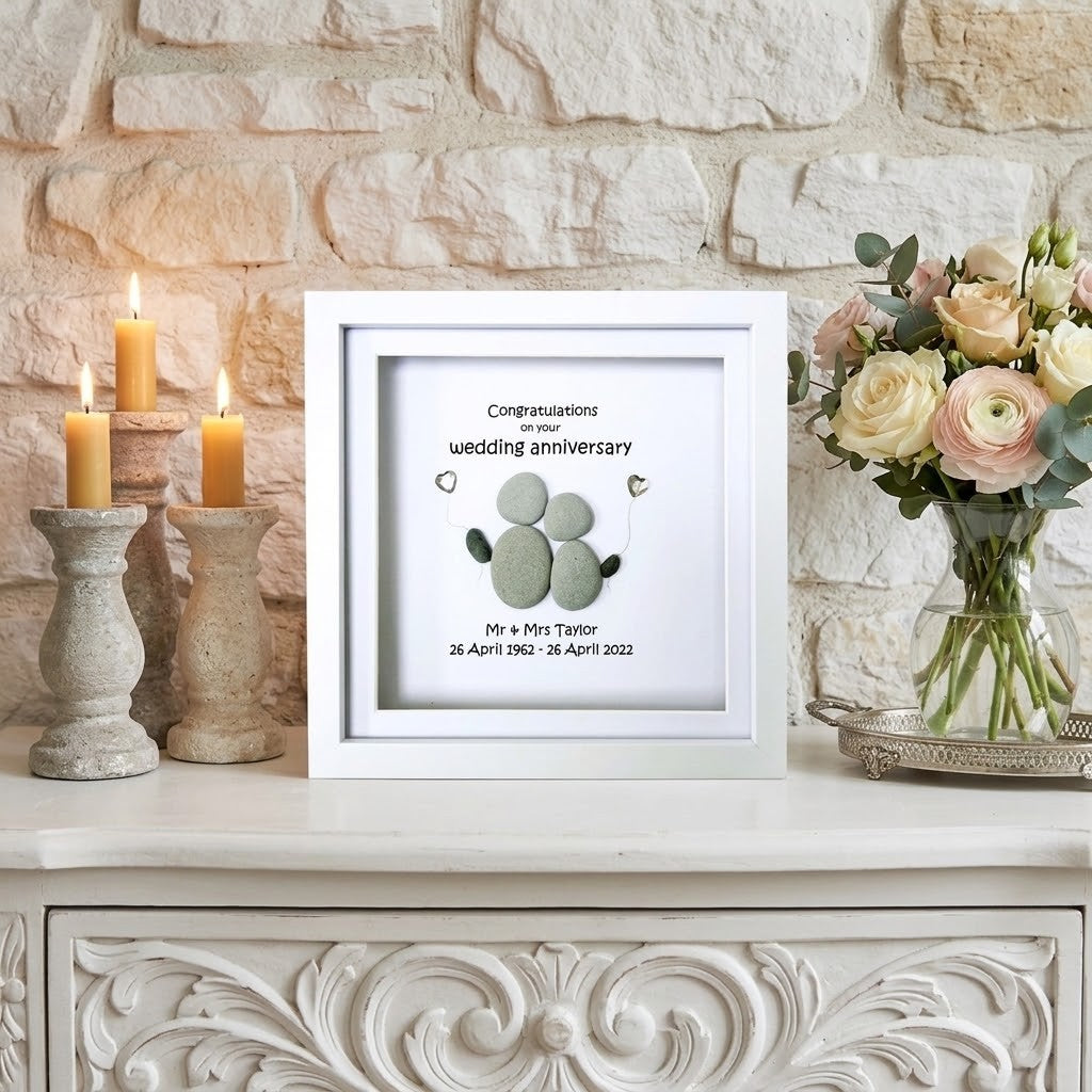 A white square framed wedding anniversary pebble art displayed on a white carved cabinet against a stone wall. The print features a green stone footprint design with the text 'Congratulations on your wedding anniversary' and personalised names and dates.