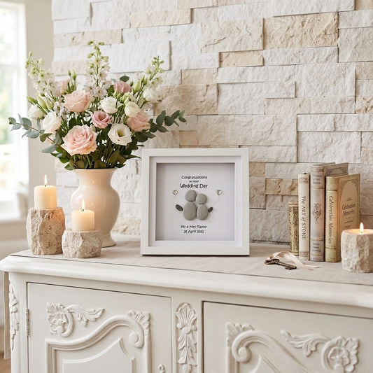 A white square framed keepsake wedding day print displayed on a white carved console table, styled with a vase of pink and white flowers, candles, and books against a light stone wall.