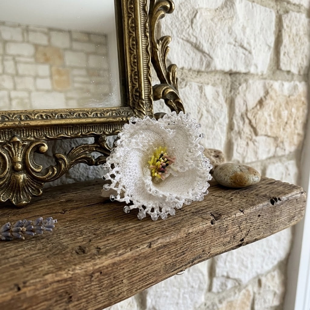 A white crochet lace flower brooch with a yellow and pink centre, displayed on a rustic wooden mantel next to a decorative gold mirror frame against a light stone wall.