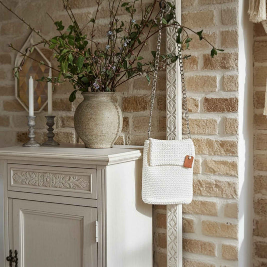 White textured handbag hanging on a decorative stand against a brick wall with a vase of branches.