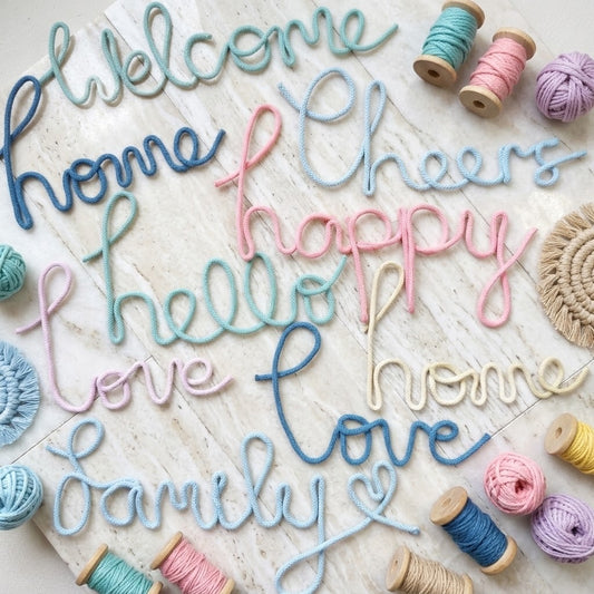 A decorative wall sign made of colourful embroidered script letters spelling out words like 'Welcome', 'home', 'Cheers', 'happy', 'hello', 'love', and 'family'. The sign is arranged on a light wood surface with yarn balls and spools of thread around it.