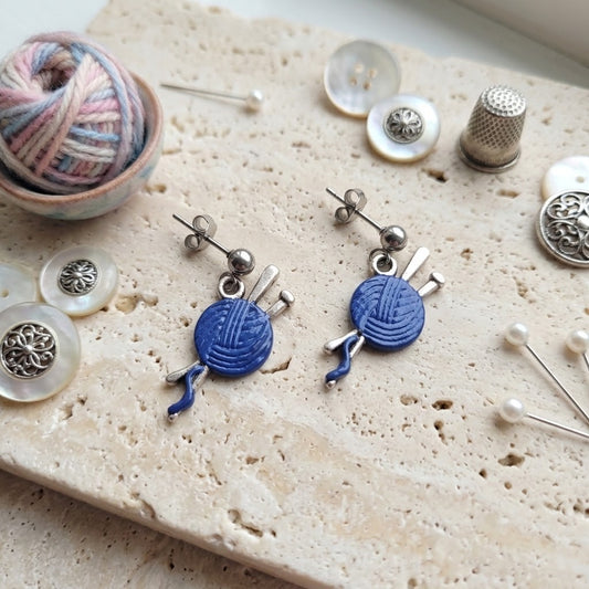 A pair of silver-toned dangle earrings shaped like blue yarn balls with knitting needles, displayed on a textured beige stone surface with sewing and crafting props including buttons, pins, a thimble, and yarn.