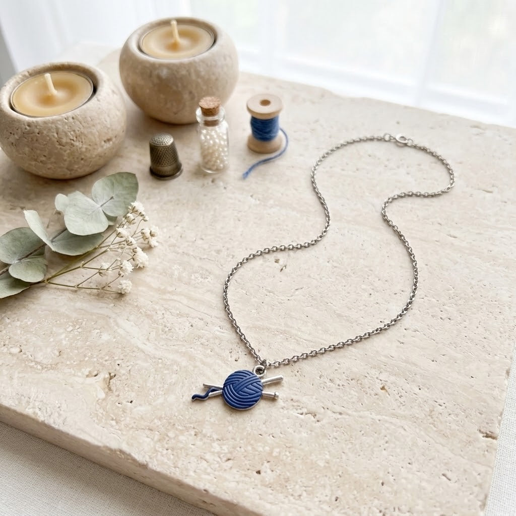 A silver-toned chain necklace with a blue and white yarn ball pendant, styled on a light stone surface with candles, eucalyptus leaves, and small spools in the background.