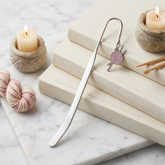 A silver metal bookmark shaped like knitting needles with a small yarn ball charm, resting on stacked cream-coloured books on a white marble surface with candles and yarn skeins as props.