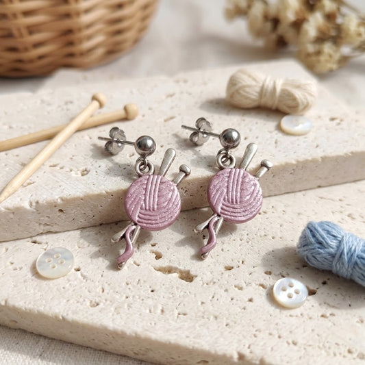 A pair of silver-toned dangle earrings with pink yarn ball charms, displayed on a textured beige stone slab with knitting props.