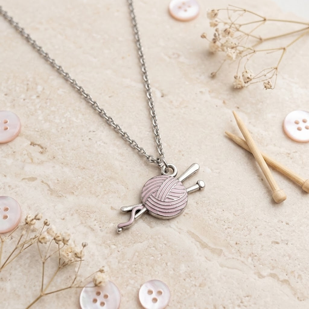 A silver chain necklace with a charm shaped like a pink and white ball of yarn wrapped around knitting needles, displayed on a textured beige surface with decorative buttons, wooden sticks, and dried flowers.