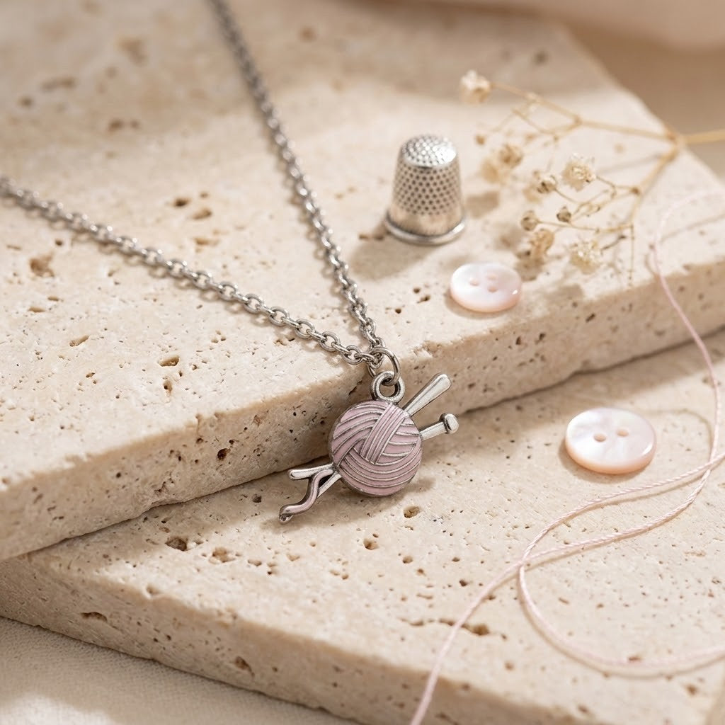 A silver-toned chain necklace with a pendant shaped like a pink ball of yarn next to knitting needles, styled on beige stone blocks with sewing-themed props including a thimble, buttons, and thread.