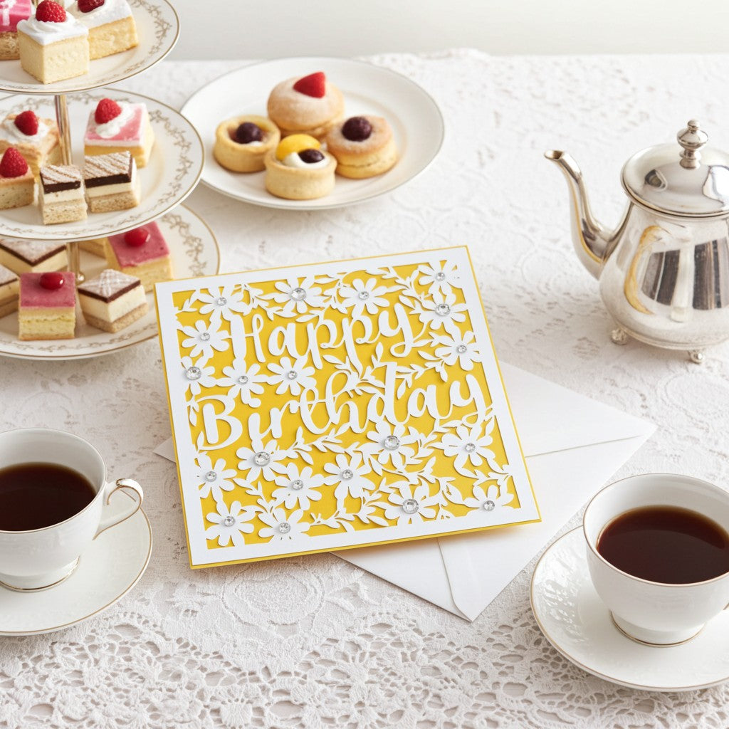 A yellow laser cut birthday card with happy birthday and flowers on a table with envelope surrounded by pretty cakes, a teapot and cups of tea.