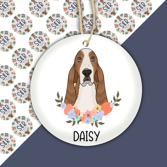 A ceramic Christmas decoration featuring an illustration of a Basset Hound dog with a floral wreath around its neck, on a white background, with the name 'Daisy' printed at the bottom.