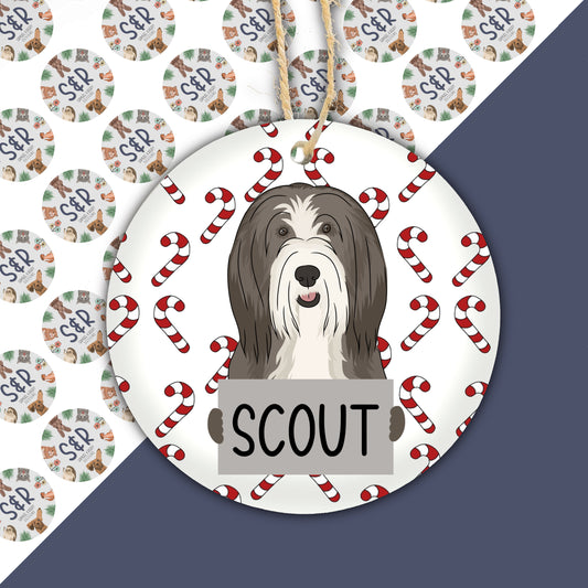 A ceramic Christmas ornament featuring a cartoon illustration of a bearded collie dog with the word 'Scout' personalised on it.