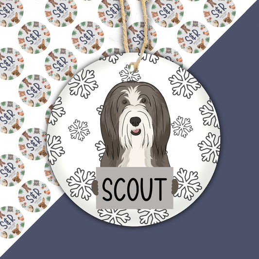A ceramic Christmas decoration featuring an illustration of a bearded collie dog with a snowflake pattern in the background.
