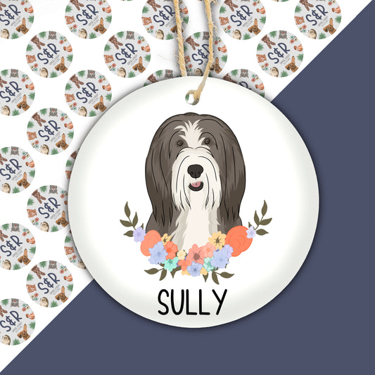 A ceramic Christmas decoration featuring an illustration of a bearded collie dog with a floral wreath background, with the name 'Sully' printed at the bottom.