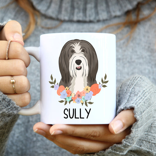 A person holding a white coffee mug with a colourful floral design and an illustration of a bearded collie dog on the side.