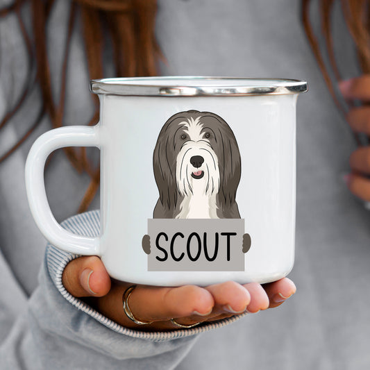 A hand holding a white enamel mug with an illustration of a Bearded Collie dog and the name 'SCOUT' printed on the front.