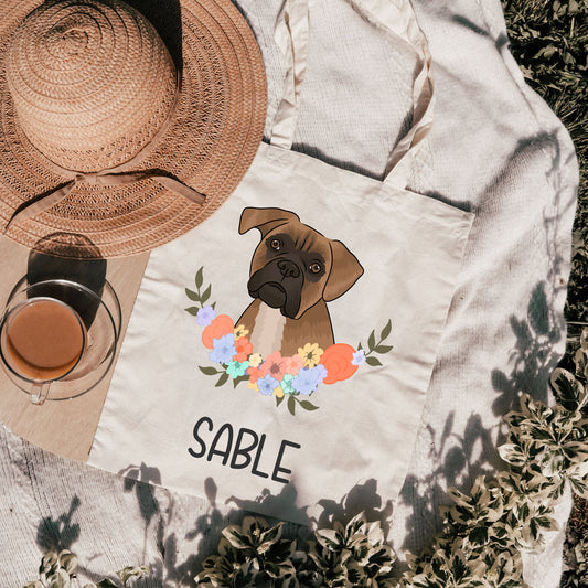 A personalised tote bag with an illustration of a dog, specifically a Boxer, surrounded by a floral pattern on a beige background. The text 'SABLE' is also visible on the bag.
