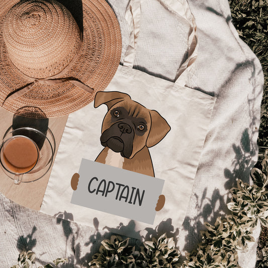 A white cotton tote bag with a printed graphic of a Boxer dog and the text 'CAPTAIN' on a brown background, with a straw hat and a glass of a beverage visible in the background.