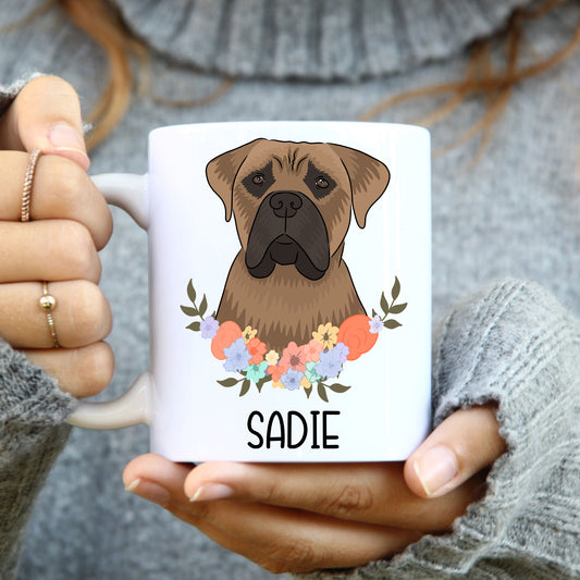 A person holding a white coffee mug with a colorful floral design and an illustration of a Bullmastiff dog on the side.