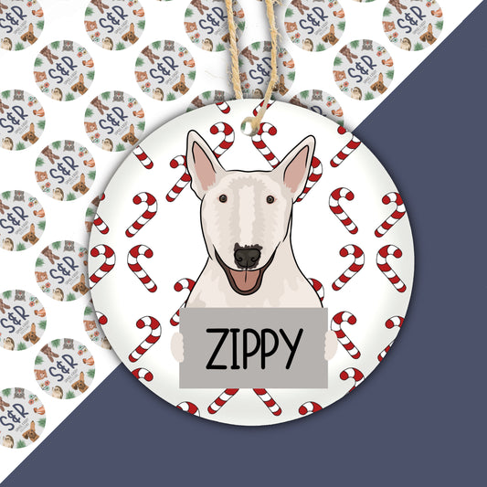 A ceramic Christmas ornament featuring a cartoon illustration of a Bull Terrier dog with the name 'Zippy' on it, surrounded by Christmas-themed decorations like trees and Santa Claus.
