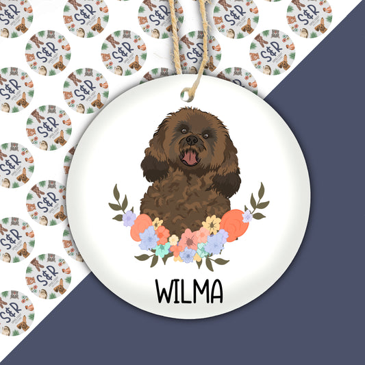 A ceramic Christmas decoration featuring an illustration of a cavapoo dog with a floral wreath background, with the name 'WILMA' at the bottom.