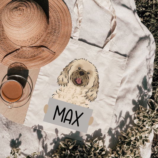 A white cotton tote bag with a personalized print of a cavachon dog named 'Max' on the front, with a green field and a straw hat in the background.