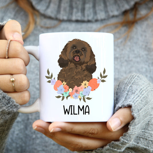 A person holding a white coffee mug with a colourful illustration of a cavapoo dog on both sides, with the name 'Wilma' printed on the mug.