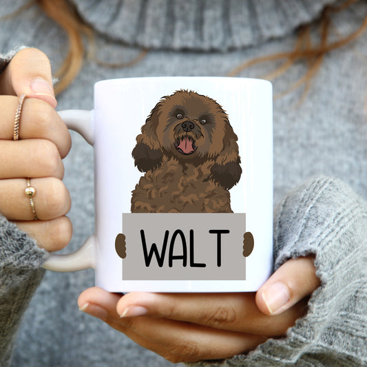 A person holding a white coffee mug with a printed image of a cavapoo dog on it.